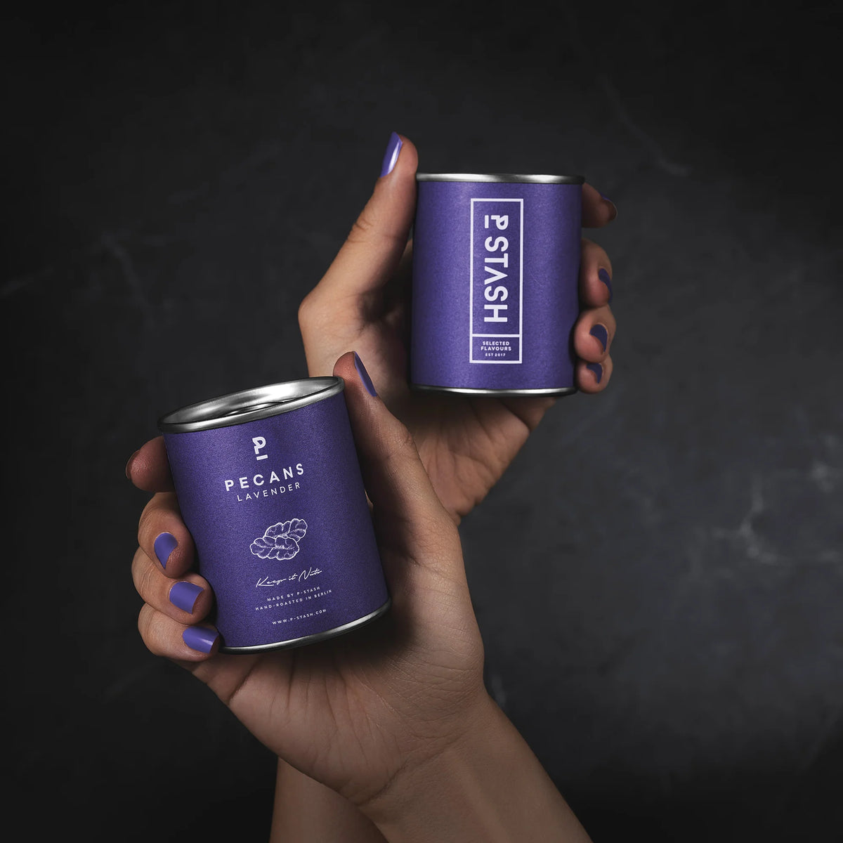 Hände mit lila lackierten Nägeln halten zwei lila P-Stash Dosen der Sorte „Pecans Lavender“ vor einem dunklen Hintergrund.