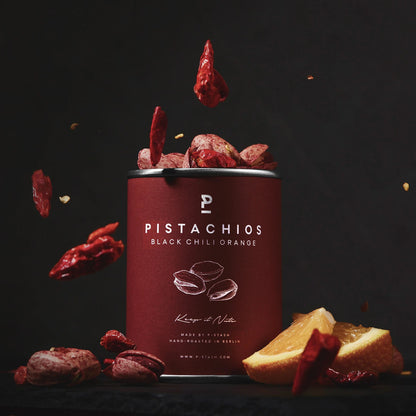 Dose mit P-Stash Pistazien, Sorte Black Chili Orange, umgeben von getrockneten Chilischoten und Orangenscheiben