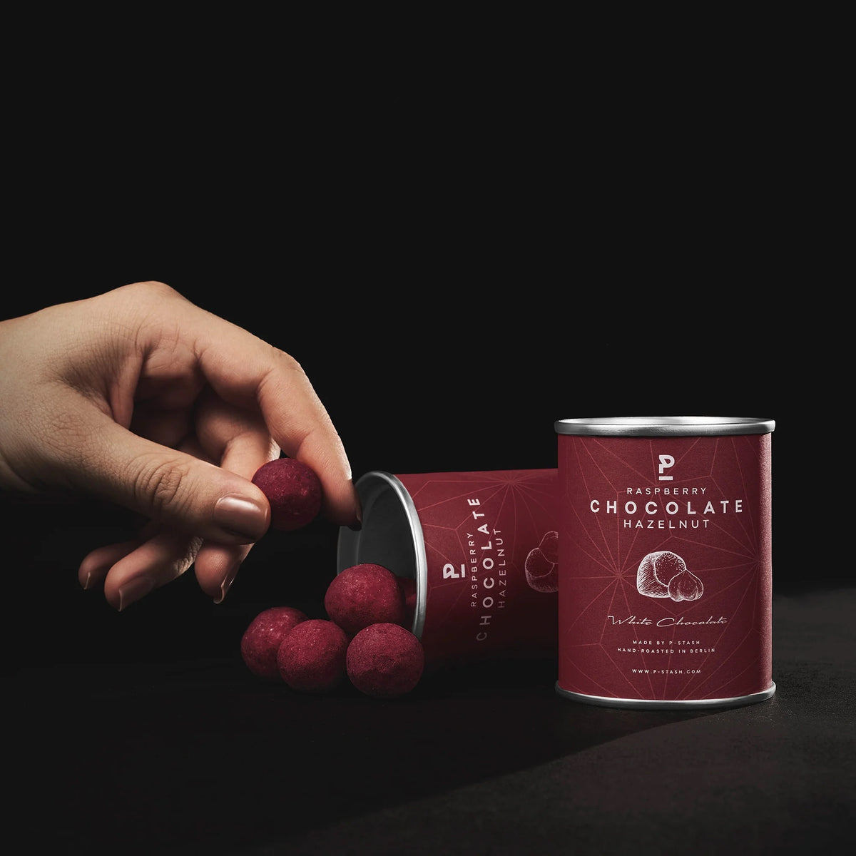 Eine aufrechte und eine liegende beerenrote P-Stash Dose mit rosa-roten Himbeer-Schoko-Haselnüssen, von denen eine nach einer Hand greift.