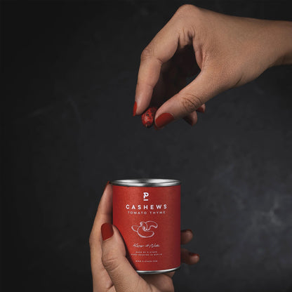 Hand mit roten Nägeln hält eine Dose Cashews mit Tomate-Thymian-Geschmack, andere Hand hält eine Cashewnuss