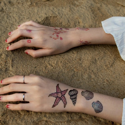 Zwei Unterarme mit temporären Tattoos von Seesternen, Muscheln und Blumen, liegend auf Sand