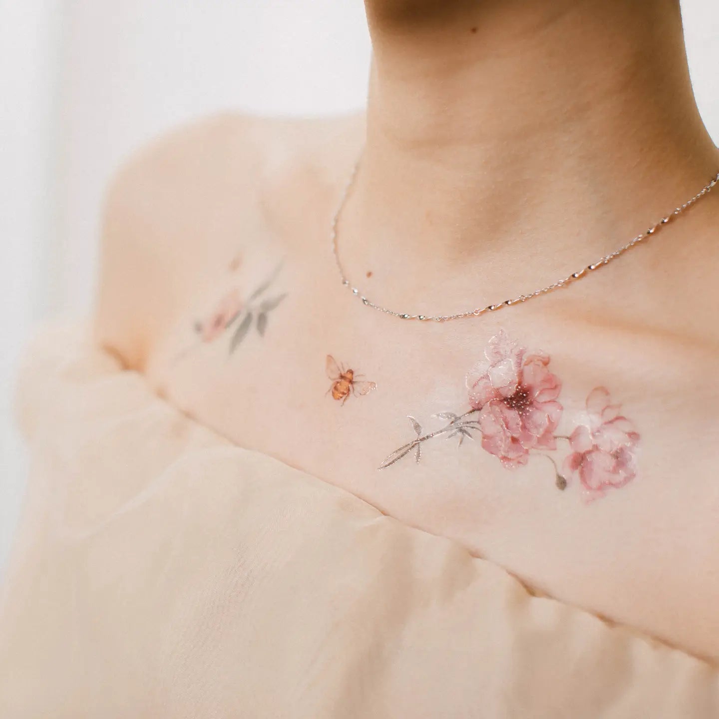 Schulter und Schlüsselbein einer Frau mit zarten Tätowierungen von rosa Blumen und einer Biene, dazu eine dünne Silberkette