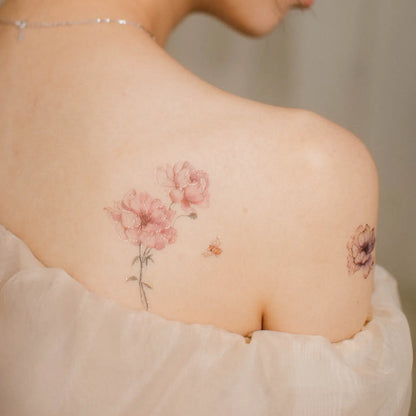 Schulter einer Frau mit zwei floralen Tattoos, eines mit rosa Blüten und einem kleinen Schmetterling, das andere mit einer dunklen Blume