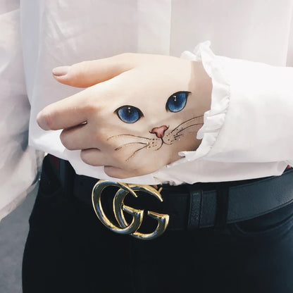 Hand mit Tattoo blauen Katzenaugen, rosa Nase und Schnurrhaaren, getragen von Person in weißer Bluse und schwarzem Gürtel mit goldener Doppelschließe