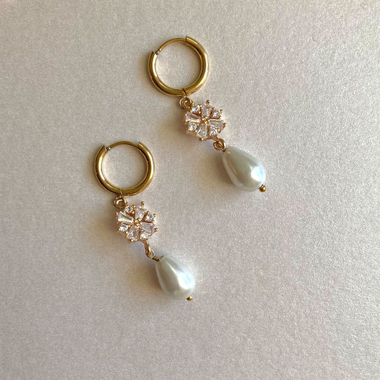 Ein Paar elegante, goldfarbene Creolen-Ohrringe auf weißem Grund. Die Ohrringe verfügen über einen abnehmbaren Anhänger, bestehend aus einem funkelnden, blumenförmigen Element aus Zirkonia-Steinen und einer darunter hängenden, tropfenförmigen weißen Perle.