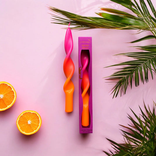 Zwei gedrehte Kerzen in Pink und Orange, eine in einer pinken Verpackung, umgeben von Orangenscheiben und grünen Palmblättern auf rosa Untergrund