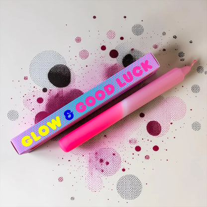 Eine pinke Dip-Dye-Stabkerze von Pink Stories mit sanftem Farbverlauf von hellem Neon-Rosa zu Altrosa, daneben liegt die passende bedruckte Geschenkbox mit der Aufschrift „GLOW & GOOD LUCK“.