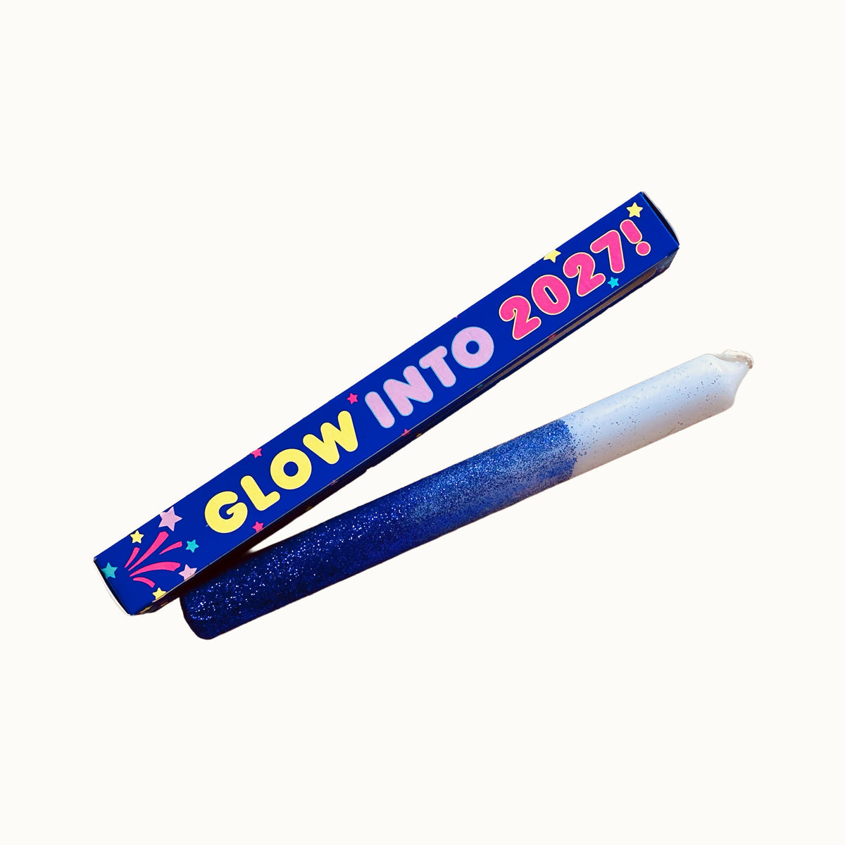 Eine Stabkerze, die zur Hälfte blau glitzert und zur Hälfte weiß ist, liegt schräg vor einer blauen Geschenkverpackung. Auf der Verpackung steht in bunter, verspielter Schrift „GLOW INTO 2027!“, verziert mit kleinen Sternen und einem Feuerwerksmotiv. Alles ist vor einem neutralen, cremefarbenen Hintergrund freigestellt.