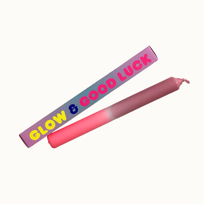 Eine pinke Dip-Dye-Stabkerze von Pink Stories mit sanftem Farbverlauf von hellem Neon-Rosa zu Altrosa, daneben liegt die passende bedruckte Geschenkbox mit der Aufschrift „GLOW & GOOD LUCK“.