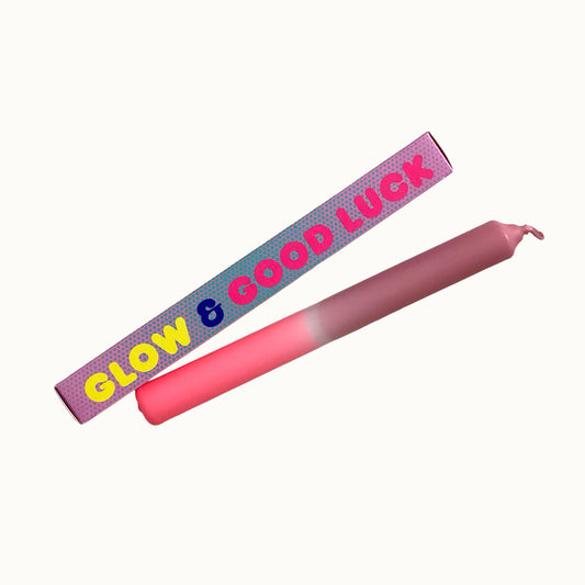 Eine pinke Dip-Dye-Stabkerze von Pink Stories mit sanftem Farbverlauf von hellem Neon-Rosa zu Altrosa, daneben liegt die passende bedruckte Geschenkbox mit der Aufschrift „GLOW & GOOD LUCK“.