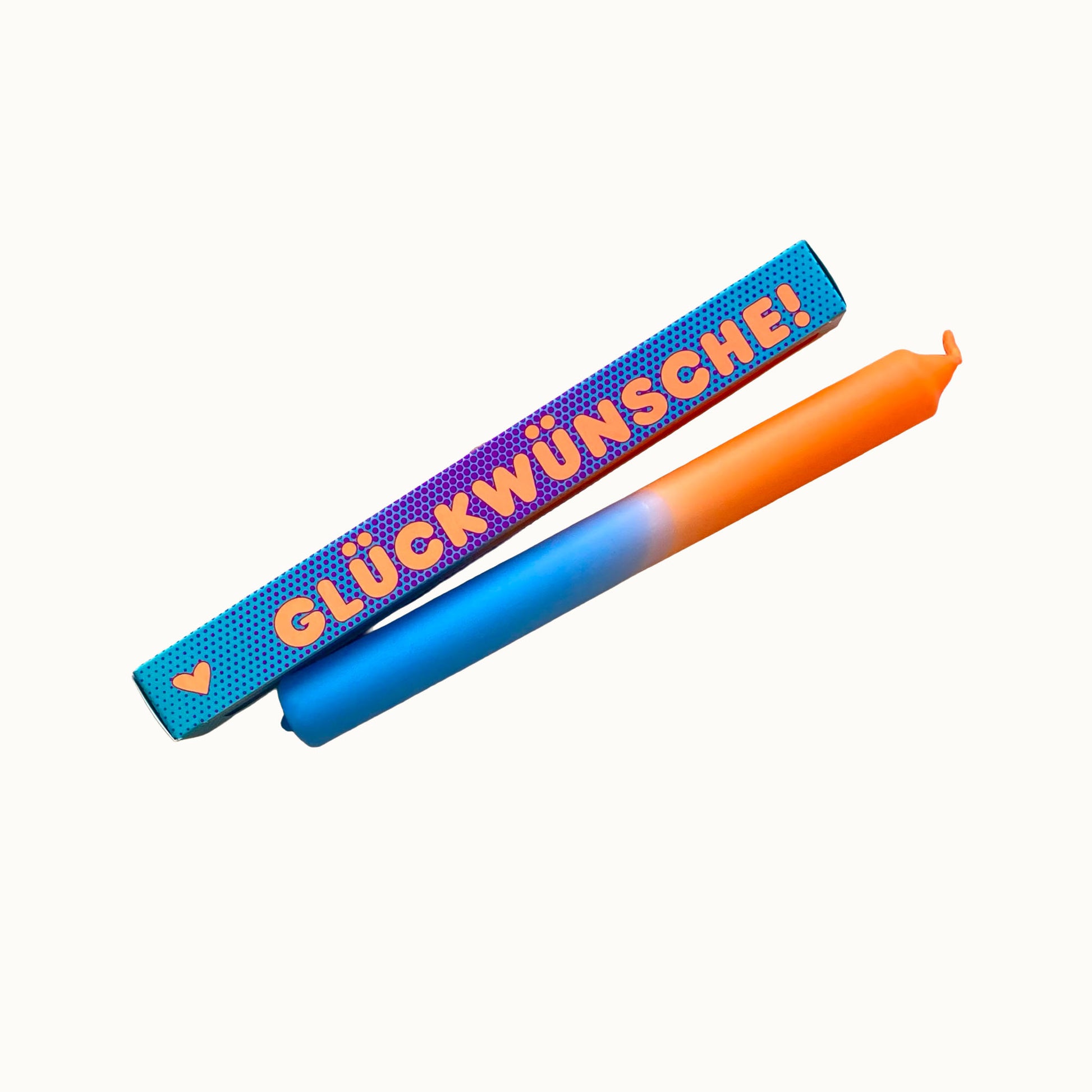 Eine zweifarbige Stabkerze in Hellblau und Orange liegt schräg vor einer türkisfarbenen Geschenkverpackung mit feinem Punktmuster. Auf der Verpackung steht in orangefarbener Schrift „GLÜCKWÜNSCHE!“, links daneben ist ein kleines orangefarbenes Herz abgebildet. Alles ist vor einem neutralen, cremefarbenen Hintergrund freigestellt.