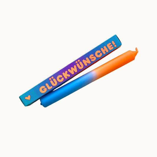 Eine zweifarbige Stabkerze in Hellblau und Orange liegt schräg vor einer türkisfarbenen Geschenkverpackung mit feinem Punktmuster. Auf der Verpackung steht in orangefarbener Schrift „GLÜCKWÜNSCHE!“, links daneben ist ein kleines orangefarbenes Herz abgebildet. Alles ist vor einem neutralen, cremefarbenen Hintergrund freigestellt.