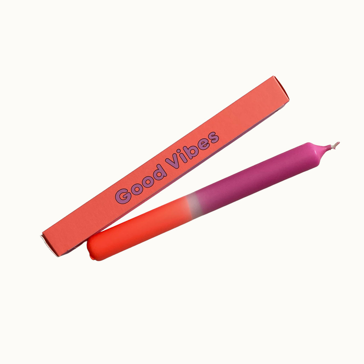 Eine Dip-Dye-Stabkerze von Pink Stories mit einem Farbverlauf von Pfirsich-Orange zu kräftigem Beere, neben der gleichfarbigen Geschenkbox mit der Aufschrift „Good Vibes“.