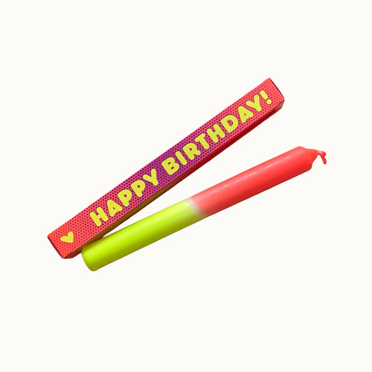 Eine zweifarbige Stabkerze in Neongelb und Neonrot liegt schräg vor einer roten Geschenkverpackung mit feinem Punktmuster. Auf der Verpackung steht in gelber Schrift „HAPPY BIRTHDAY!“, links daneben ist ein kleines gelbes Herz abgebildet. Alles ist vor einem neutralen, cremefarbenen Hintergrund freigestellt.