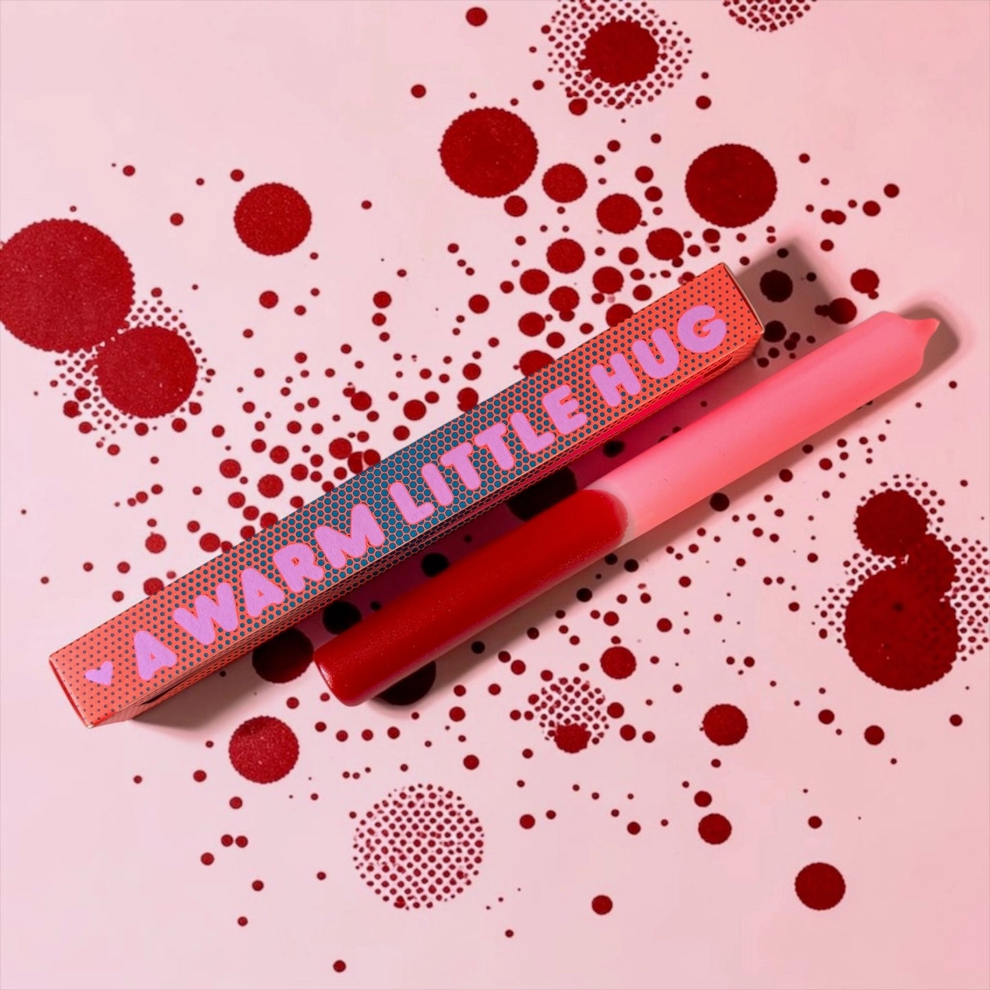 Zweifarbige Stabkerze in Rot und Rosa auf einem rosa Hintergrund mit roten Punkten. Daneben liegt die rot gepunktete Schachtel mit der rosa Aufschrift „A WARM LITTLE HUG“.