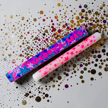 Pink Stories Stabkerze Merry X-Mas in Weiß und Rosa mit pinken Sternen neben bunter Geschenkverpackung auf Konfetti-Hintergrund
