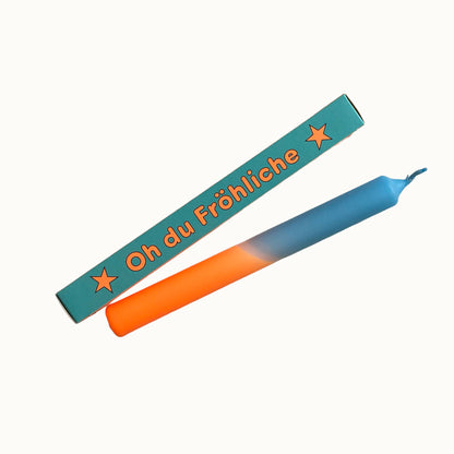 Eine weihnachtliche Dip-Dye-Stabkerze in Orange und Himmelblau von Pink Stories, neben der hellblauen Geschenkbox mit der Aufschrift „Oh du Fröhliche".