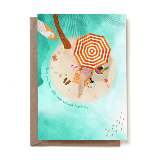Eine illustrierte Geburtstagskarte im sommerlichen Urlaubsdesign mit der geschwungenen Aufschrift „May you have the most relaxed birthday!“. Die Szene zeigt eine Luftansicht einer kleinen, runden Sandinsel inmitten von türkisfarbenem Wasser. Unter einer Palme und einem rot-weiß gestreiften Sonnenschirm liegt eine Frau im Bikini entspannt auf einer Decke. Neben ihr befinden sich ein fruchtiger Cocktail, eine Obstschale mit Wassermelone, Flip-Flops, Muscheln, Seesterne und ein aufgeschlagenes Buch. Im Wasser 