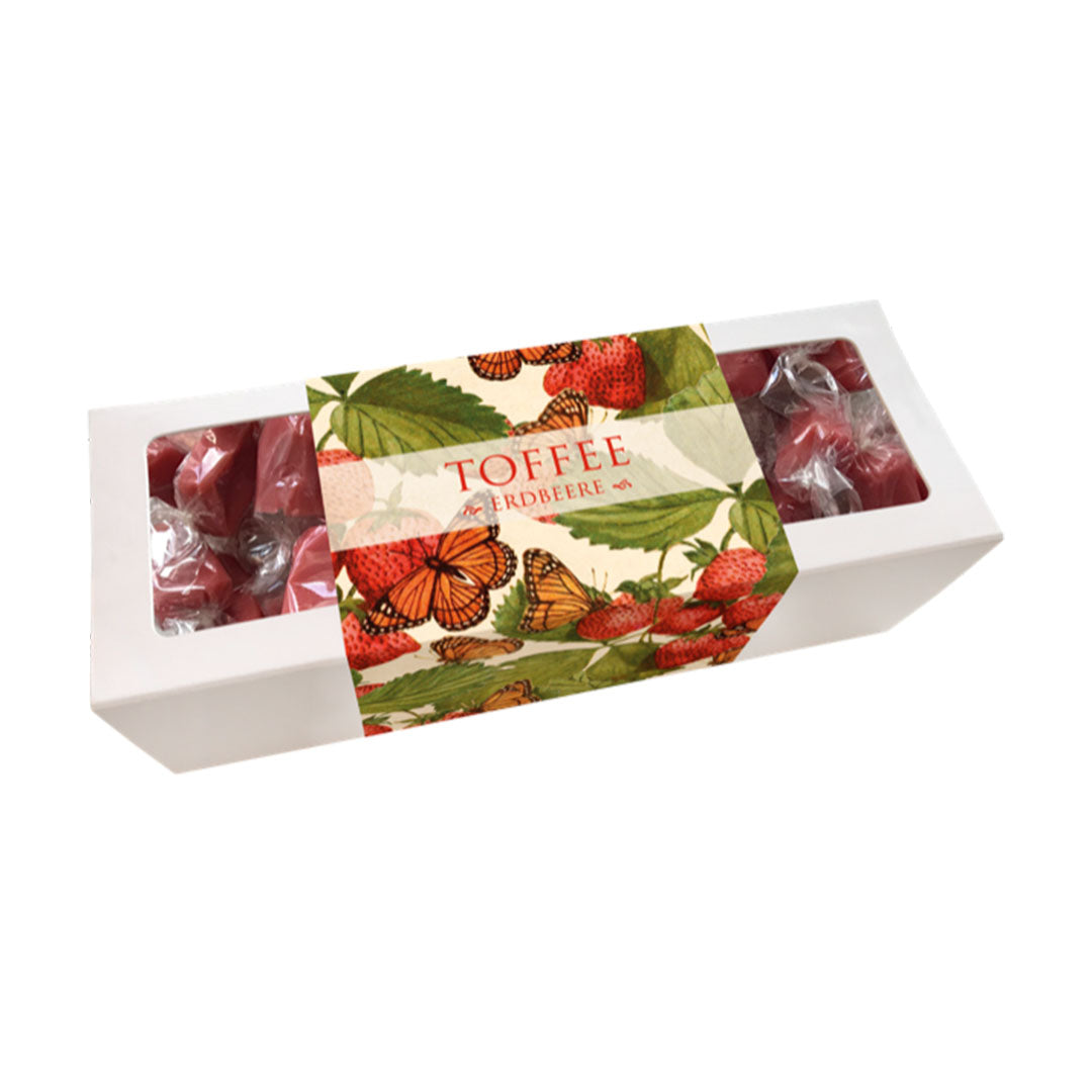 Eine weiße längliche Box mit Erdbeer-Toffees. Das Design der Banderole zeigt ein nostalgisches Naturmotiv mit roten Erdbeeren und orangefarbenen Schmetterlingen (Monarchfalter). Im Sichtfenster liegen rötliche, in Klarsichtfolie gewickelte Toffee-Bonbons. Titel: „TOFFEE ERDBEERE“.