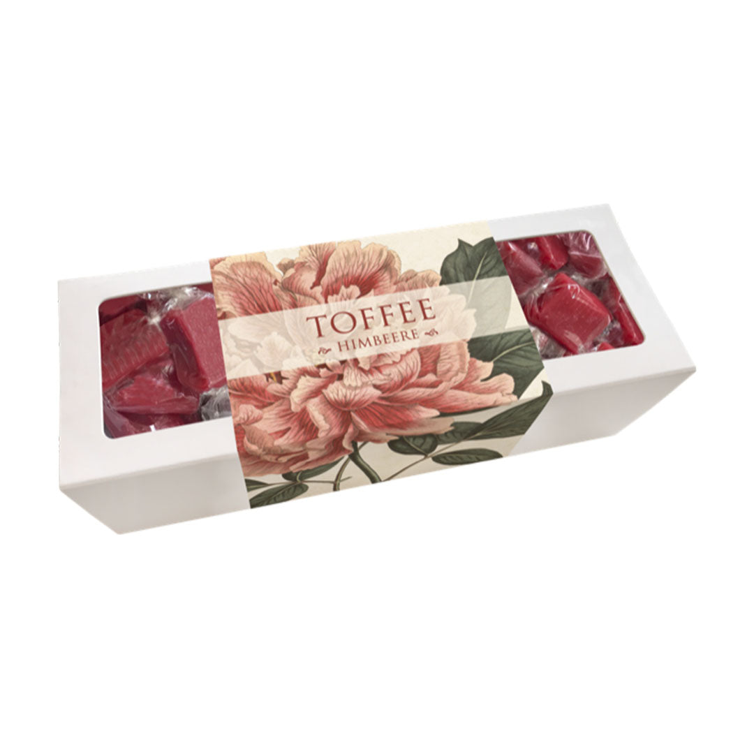 Weiße Geschenkverpackung der Marke Sköna Ting für Himbeer-Toffees. Die Banderole besticht durch eine große, detaillierte Illustration einer rosa Pfingstrose im botanischen Stil. Die Box enthält dunkelrosa Toffees, die durch das Fenster links und rechts neben der Banderole erkennbar sind. Aufschrift: „TOFFEE HIMBEERE“.