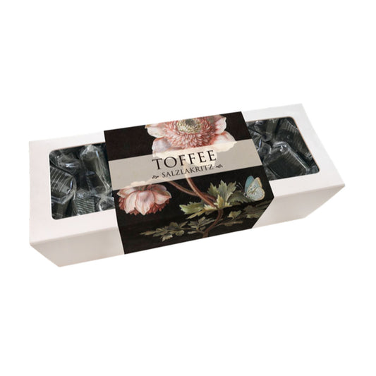 Weiße rechteckige Verpackung mit durchsichtiger Folie, schwarzem Etikett mit Blumenmotiv und Schriftzug 'TOFFEE mit SALZLAKRITZ'.