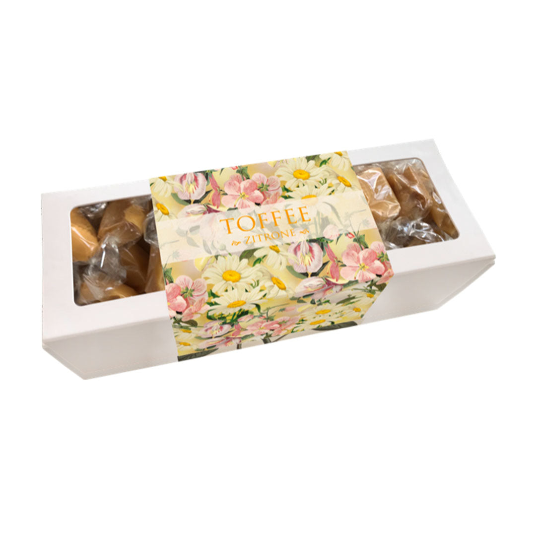 Eine weiße Geschenkbox mit Sichtfenster, gefüllt mit einzeln eingewickelten Zitronen-Toffees. Die Banderole zeigt eine botanische Illustration von weißen Gänseblümchen und zartrosa Blüten auf gelbem Grund mit der Aufschrift „TOFFEE ZITRONE“.