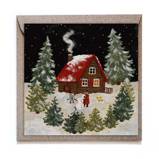 Quadratische Studio Satsch Winterkarte: Eine eingeschneite Blockhütte mit rotem Dach im Tannenwald bei Nacht. Im Vordergrund bauen zwei Personen im Schnee einen Schneemann.