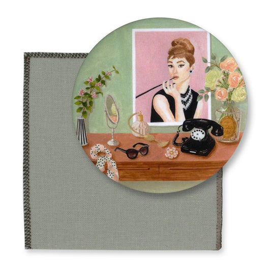 Runder Taschenspiegel mit Illustration im Audrey Hepburn Stil: Eine Frau mit Hochsteckfrisur und Perlenkette blickt über einen Schreibtisch mit schwarzem Wählscheibentelefon, Sonnenbrille und Blumenmaske.