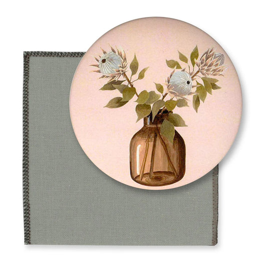 Runder Taschenspiegel mit einer botanischen Illustration von zwei Protea-Blumen in einer braunen Glasvase auf einem hellrosa Hintergrund. Im Hintergrund ist ein Teil einer  salbeigrünen  Schutzhülle zu sehen.