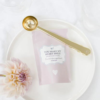 Pinke Teeverpackung „YOU MAKE MY HEART SMILE“ auf einem weißen Teller, dekoriert mit einer passenden Blüte und einem goldenen Dosierlöffel