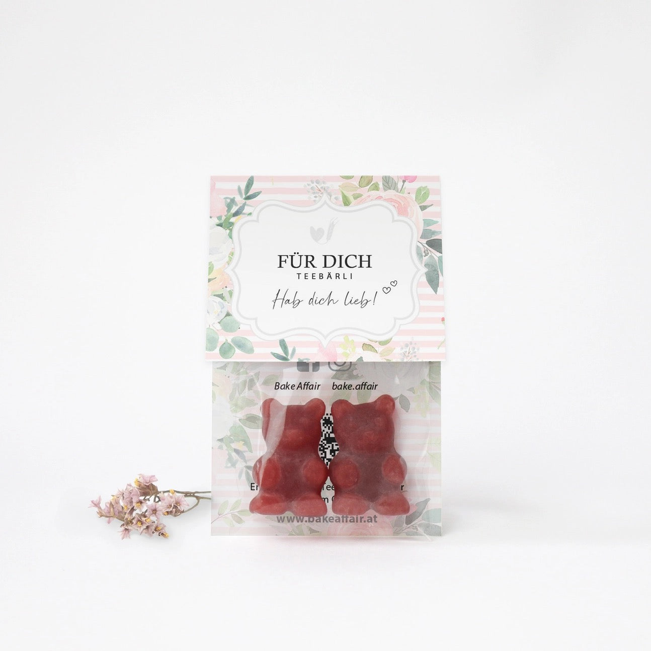 Zwei rote Gummibärchen in transparenter Verpackung mit Aufkleber 'FÜR DICH TEEBÄRLI Hab dich lieb!' und getrocknete Blüten daneben.