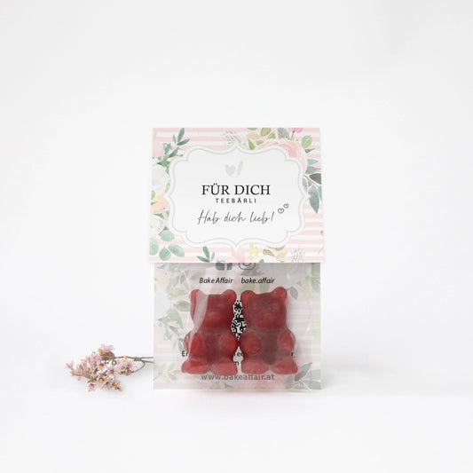Zwei rote Gummibärchen in transparenter Verpackung mit Aufkleber 'FÜR DICH TEEBÄRLI Hab dich lieb!' und getrocknete Blüten daneben.