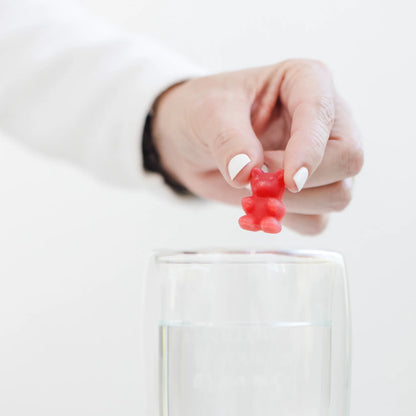 Hand mit weiß lackierten Fingernägeln hält roten Gummibär über ein Glas mit klarem Wasser.