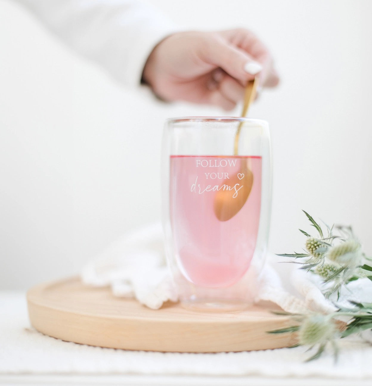 Glas mit rosa Flüssigkeit und Aufschrift 'FOLLOW YOUR dreams', daneben weiße Blüten, Hand rührt mit goldfarbenem Löffel.