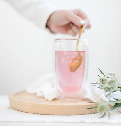 Glas mit rosa Flüssigkeit und Aufschrift 'FOLLOW YOUR dreams', daneben weiße Blüten, Hand rührt mit goldfarbenem Löffel.