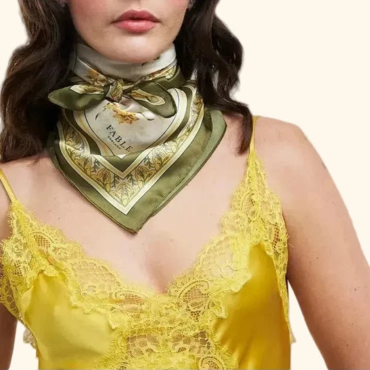 Frau trägt das Fable England Tuch als modisches Halstuch geknotet. Das Tuch hat einen olivgrünen Rand und gelbe Blumenmuster, kombiniert mit einem gelben Spitzenoberteil.