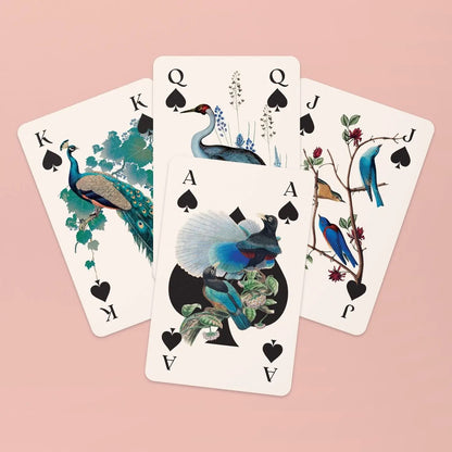 Vier ausgewählte Spielkarten der Pik-Familie sind fächerförmig auf einem zartrosa Hintergrund angeordnet. Zu sehen sind der König mit einem Pfau, die Dame mit einem eleganten grauen Kranich vor blauen Blumen, der Bube mit blau-gelben Vögeln auf einem Ast und im Vordergrund das Pik-As, dessen großes schwarzes Symbol kunstvoll mit zwei Paradiesvögeln illustriert ist.
