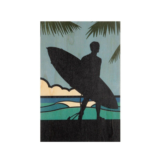 Eine stimmungsvolle Postkarte auf Holz, die die dunkle Silhouette eines Surfers mit Brett am Strand zeigt. Im Hintergrund sind Palmenblätter, Wellen und ein sanfter blauer Himmel zu sehen.