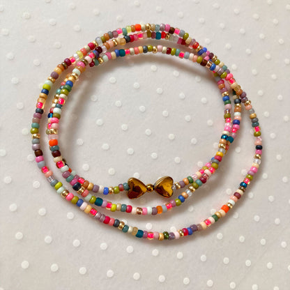 Mehrreihiges Wickelarmband aus präzisen, bunten Miyuki-Glasperlen in verschiedenen Farben wie Türkis, Pink und Gelb. In der Mitte befindet sich ein glänzender Anhänger aus zwei goldfarbenen Herzen aus Hämatit-Stein. Der Schmuck liegt auf einem neutralen, hellen Hintergrund.