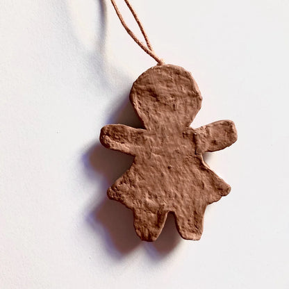Unbemalter, unfertiger Lebkuchenfrau-Anhänger aus Pappmaché an einer beigen Schnur, aufgenommen vor einem weißen Hintergrund. Der Anhänger hat eine raue, braune Oberfläche und eine flache Rückseite.