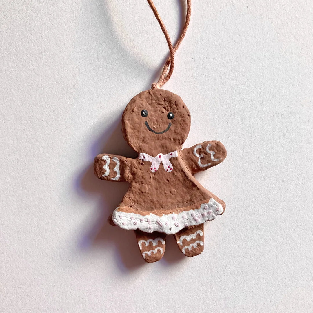Ein vollständig bemaltes Lebkuchenfrau-Anhänger aus Pappmaché an einer beigen Schnur, vor einem weißen Hintergrund. Die Figur hat ein gemaltes Gesicht, eine rosa Schleife um den Hals, ein rosa Kleid mit weißem, gepunktetem Rocksaum und weiße Schnörkel an den Armen und Beinen.