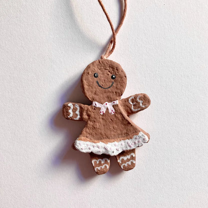 Ein vollständig bemaltes Lebkuchenfrau-Anhänger aus Pappmaché an einer beigen Schnur, vor einem weißen Hintergrund. Die Figur hat ein gemaltes Gesicht, eine rosa Schleife um den Hals, ein rosa Kleid mit weißem, gepunktetem Rocksaum und weiße Schnörkel an den Armen und Beinen.