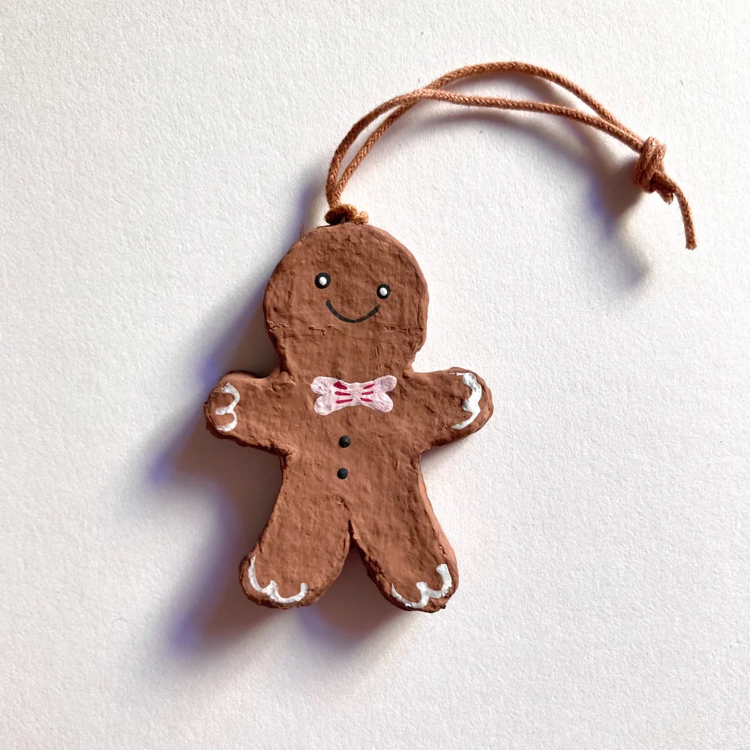 Ein vollständig bemalter Lebkuchenmann-Anhänger aus Pappmaché an einer beigen Schnur, vor einem weißen Hintergrund. Die Figur hat ein lächelndes Gesicht, eine rosa Fliege, zwei schwarze Knöpfe auf der Brust und weiße Schnörkel an den Händen und Füßen.