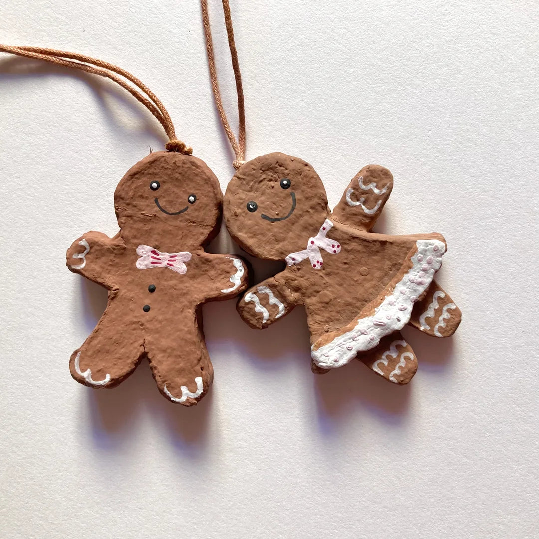 Ein Paar bemalte Lebkuchenmann- und Lebkuchenfrau-Anhänger aus Pappmaché an beigen Schnüren, nebeneinander liegend vor einem weißen Hintergrund. Der Lebkuchenmann trägt eine rosa Fliege und schwarze Knöpfe. Die Lebkuchenfrau trägt ein rosa Kleid mit weißem Rocksaum und einen rosa Schal. Beide haben gemalte Gesichter und Verzierungen an den Gliedmaßen.