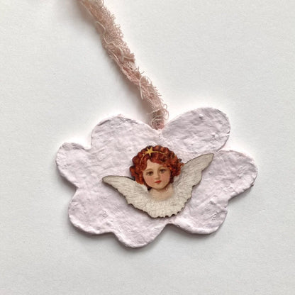 Handgefertigter Engel-Anhänger aus Pappmache auf Off-White Hintergrund mit zartrosa Fransenband.