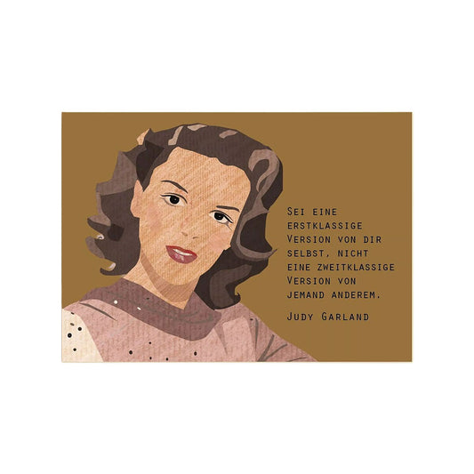 Eine im Querformat gestaltete Postkarte auf strukturierter Pappe. Die linke Hälfte zeigt eine Illustration von Judy Garland mit braunem, gewelltem Haar und einem rötlichen Oberteil vor einem braunen Hintergrund. Rechts daneben steht das Zitat: „Sei eine erstklassige Version von dir selbst, nicht eine zweitklassige Version von jemand anderem.“ Darunter befindet sich ihr Name „Judy Garland“.