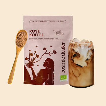 Zubereiteter Rose Koffee als Iced Latte mit Milchschaum neben der Originalverpackung und einem Holzlöffel mit dem dunklen Instant-Pulver.
